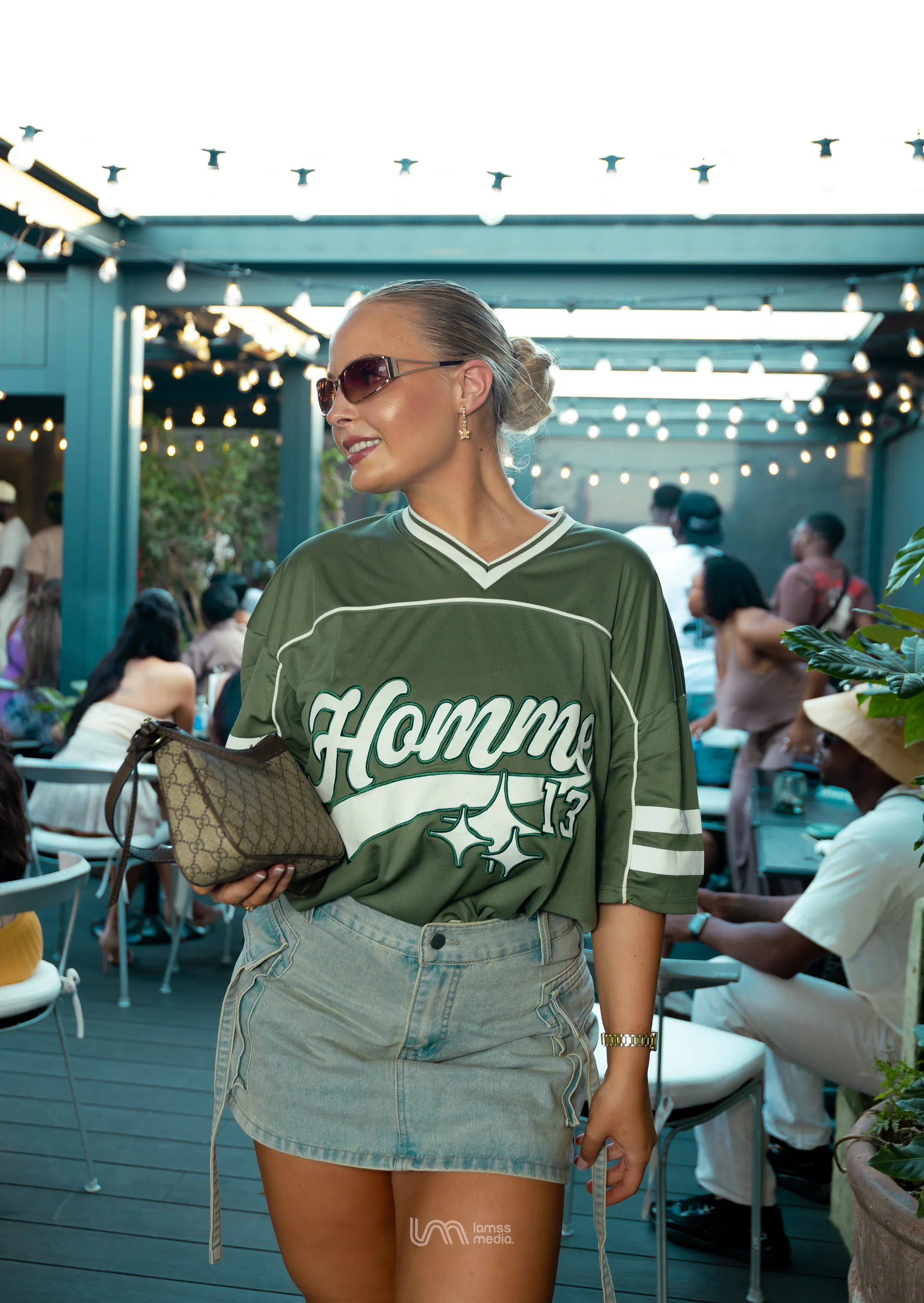 A Woman In A Green Jersey And Denim Skirt Looking To The Side At An Outdoor Patio With String Lights.